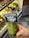 A hand holding a packaged dill pickle in front of a refrigerator stocked with various bottled drinks. The packaging is clear with a blue label and features a cartoon image of a pickle.