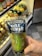 A hand holding a packaged dill pickle in front of a refrigerator stocked with various bottled drinks. The packaging is clear with a blue label and features a cartoon image of a pickle.