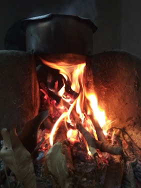 A traditional wood-burning stove with a metal pot resting on top. Flames and embers can be seen underneath, creating a warm glow. The fire is surrounded by stones or a protective barrier.
