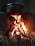 A traditional wood-burning stove with a metal pot resting on top. Flames and embers can be seen underneath, creating a warm glow. The fire is surrounded by stones or a protective barrier.