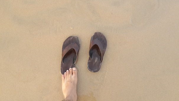 A pair of brown flip-flops rests on wet sand with a foot next to them. The sand appears smooth with a slightly darker tone near the water.