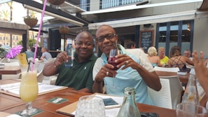 Two people are sitting at an outdoor restaurant enjoying drinks. The atmosphere is casual and relaxed, with other patrons visible in the background. The table is set with menus and drinks, including a colorful tropical cocktail with a straw and an umbrella. The setting appears to be warm and summery, with people wearing light clothing.