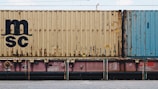 A close-up view of a freight train with stacked shipping containers. The upper container has a beige color with noticeable signs of rust and a large 'MSC' logo. The container below is partially visible and has a pinkish color. A blue container is seen adjacent to the beige one. The containers and the train feature some industrial details like labels and signs.