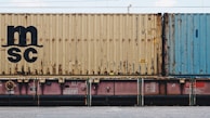 A close-up view of a freight train with stacked shipping containers. The upper container has a beige color with noticeable signs of rust and a large 'MSC' logo. The container below is partially visible and has a pinkish color. A blue container is seen adjacent to the beige one. The containers and the train feature some industrial details like labels and signs.