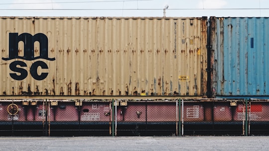 A close-up view of a freight train with stacked shipping containers. The upper container has a beige color with noticeable signs of rust and a large 'MSC' logo. The container below is partially visible and has a pinkish color. A blue container is seen adjacent to the beige one. The containers and the train feature some industrial details like labels and signs.