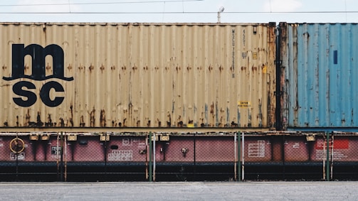 A close-up view of a freight train with stacked shipping containers. The upper container has a beige color with noticeable signs of rust and a large 'MSC' logo. The container below is partially visible and has a pinkish color. A blue container is seen adjacent to the beige one. The containers and the train feature some industrial details like labels and signs.