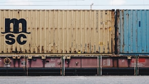 A close-up view of a freight train with stacked shipping containers. The upper container has a beige color with noticeable signs of rust and a large 'MSC' logo. The container below is partially visible and has a pinkish color. A blue container is seen adjacent to the beige one. The containers and the train feature some industrial details like labels and signs.