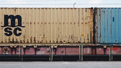 A close-up view of a freight train with stacked shipping containers. The upper container has a beige color with noticeable signs of rust and a large 'MSC' logo. The container below is partially visible and has a pinkish color. A blue container is seen adjacent to the beige one. The containers and the train feature some industrial details like labels and signs.