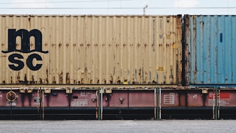 A close-up view of a freight train with stacked shipping containers. The upper container has a beige color with noticeable signs of rust and a large 'MSC' logo. The container below is partially visible and has a pinkish color. A blue container is seen adjacent to the beige one. The containers and the train feature some industrial details like labels and signs.