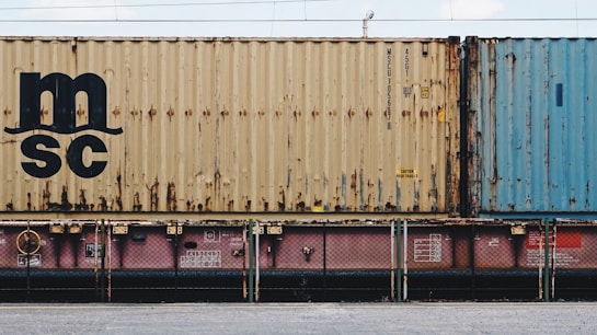 A close-up view of a freight train with stacked shipping containers. The upper container has a beige color with noticeable signs of rust and a large 'MSC' logo. The container below is partially visible and has a pinkish color. A blue container is seen adjacent to the beige one. The containers and the train feature some industrial details like labels and signs.
