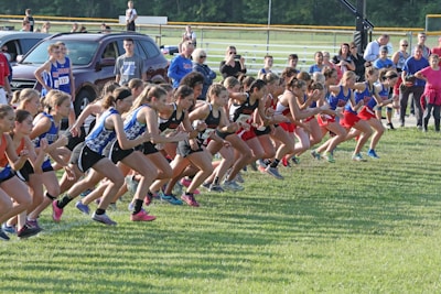 A group of female runners at the starting line of a race on a grassy field. They are dressed in various athletic uniforms, with some wearing blue and white, while others are in red and black. Spectators can be seen in the background, standing near parked cars and a fence.