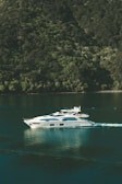 A luxurious white yacht glides smoothly over calm green waters, creating gentle ripples. It is situated near a lush, forested hillside that rises steeply from the water's edge, covered in dense green foliage.