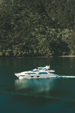 A luxurious white yacht glides smoothly over calm green waters, creating gentle ripples. It is situated near a lush, forested hillside that rises steeply from the water's edge, covered in dense green foliage.