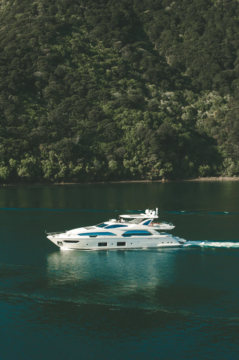 An aerial view of a white motor yacht cutting across deep green water below a forested shore.