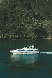 A luxurious white yacht glides smoothly over calm green waters, creating gentle ripples. It is situated near a lush, forested hillside that rises steeply from the water's edge, covered in dense green foliage.