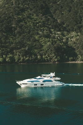 A luxurious white yacht glides smoothly over calm green waters, creating gentle ripples. It is situated near a lush, forested hillside that rises steeply from the water's edge, covered in dense green foliage.