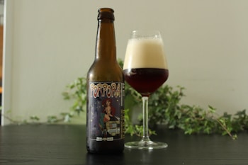 A brown beer bottle with a label that reads 'Porpora' beside a glass filled with a dark beer topped with a creamy foam. Both items are set against a muted background with green foliage.