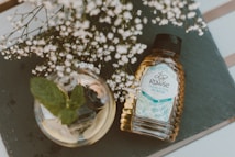 A bottle of Rowse Acacia honey lies on a slate surface, positioned beside a glass jar containing mint leaves. Delicate white flowers branch out above the honey bottle, adding a natural touch to the composition. Soft lighting enhances the earthy and inviting atmosphere of the scene.