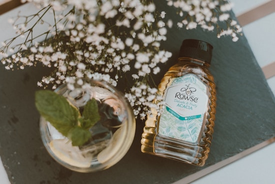 A bottle of Rowse Acacia honey lies on a slate surface, positioned beside a glass jar containing mint leaves. Delicate white flowers branch out above the honey bottle, adding a natural touch to the composition. Soft lighting enhances the earthy and inviting atmosphere of the scene.