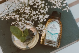 A bottle of Rowse Acacia honey lies on a slate surface, positioned beside a glass jar containing mint leaves. Delicate white flowers branch out above the honey bottle, adding a natural touch to the composition. Soft lighting enhances the earthy and inviting atmosphere of the scene.
