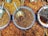 Several bowls filled with a variety of dried spices and herbs are placed in a colorful arrangement. Each bowl contains different textures and shades, with scoops resting inside, indicating they are for sale in a market setting.