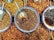 Several bowls filled with a variety of dried spices and herbs are placed in a colorful arrangement. Each bowl contains different textures and shades, with scoops resting inside, indicating they are for sale in a market setting.