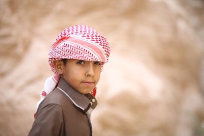 A young boy wearing traditional Middle Eastern attire with a red and white keffiyeh and a brown shirt. He stands against a blurred, neutral background, looking over his shoulder with a calm expression.