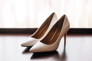 A pair of elegant beige high-heeled shoes is placed on a glossy dark surface with soft lighting creating a warm ambiance. The shoes have a textured finish and a sleek silhouette, complemented by their pointed toes and slender heels.