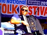 A musician wearing sunglasses plays an accordion on stage at a folk festival. The background features a large banner with the words 'Folk Festival' prominently displayed. The musician is dressed casually and appears focused on his performance.