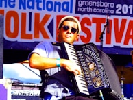 A musician wearing sunglasses plays an accordion on stage at a folk festival. The background features a large banner with the words 'Folk Festival' prominently displayed. The musician is dressed casually and appears focused on his performance.