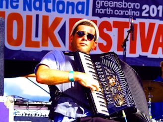 A musician wearing sunglasses plays an accordion on stage at a folk festival. The background features a large banner with the words 'Folk Festival' prominently displayed. The musician is dressed casually and appears focused on his performance.