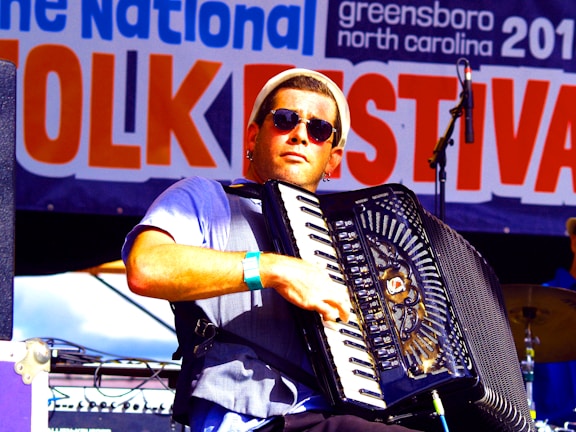 A musician wearing sunglasses plays an accordion on stage at a folk festival. The background features a large banner with the words 'Folk Festival' prominently displayed. The musician is dressed casually and appears focused on his performance.