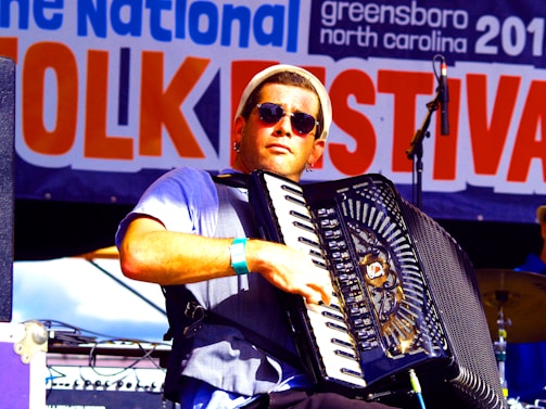 A musician wearing sunglasses plays an accordion on stage at a folk festival. The background features a large banner with the words 'Folk Festival' prominently displayed. The musician is dressed casually and appears focused on his performance.