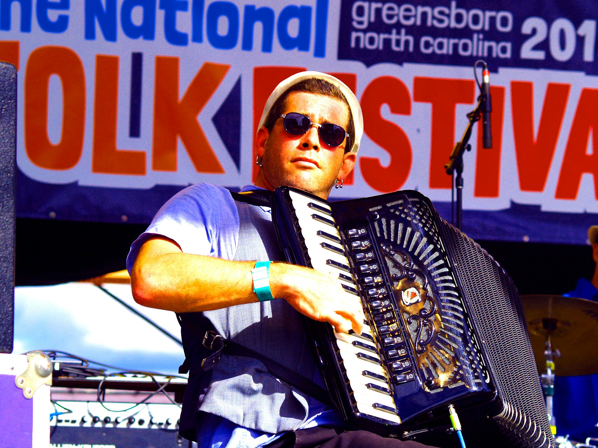 A musician wearing sunglasses plays an accordion on stage at a folk festival. The background features a large banner with the words 'Folk Festival' prominently displayed. The musician is dressed casually and appears focused on his performance.