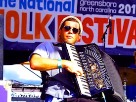 A musician wearing sunglasses plays an accordion on stage at a folk festival. The background features a large banner with the words 'Folk Festival' prominently displayed. The musician is dressed casually and appears focused on his performance.