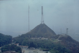 A hill is topped with a large radio or telecommunications tower, surrounded by lush greenery and smaller buildings. Additional antennas and towers can be seen in the vicinity. The landscape is hazy, suggesting fog or atmospheric conditions.
