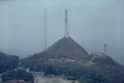 A hill is topped with a large radio or telecommunications tower, surrounded by lush greenery and smaller buildings. Additional antennas and towers can be seen in the vicinity. The landscape is hazy, suggesting fog or atmospheric conditions.