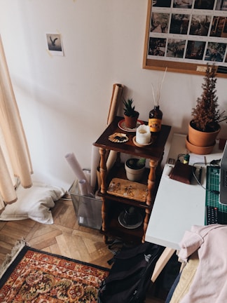 A cozy corner in a room featuring a small wooden shelf with potted plants, a candle, and a decorative bottle. A white desk with a cutting mat and a remote control is adjacent to the shelf. A section of a framed collage with photographs hangs above the shelf, and a small picture is taped to the wall. Rolled papers are stored in a wire basket on the floor. A patterned rug and a soft cushion are also visible.