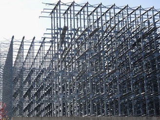 A complex structure of metal scaffolding forms an intricate geometric pattern. The grid-like arrangement of bars and beams creates a sense of depth and perspective, with the structure extending into the distance.