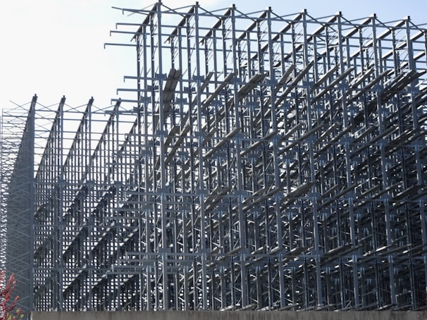 A complex structure of metal scaffolding forms an intricate geometric pattern. The grid-like arrangement of bars and beams creates a sense of depth and perspective, with the structure extending into the distance.