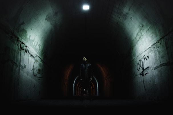 A person stands at the center of a dimly lit underground tunnel with graffiti on the concrete walls. The tunnel is illuminated by a single overhead light, and there is a sense of depth as the tunnel extends into darkness. The atmosphere is mysterious, and the lighting casts shadows that enhance the eerie ambiance.