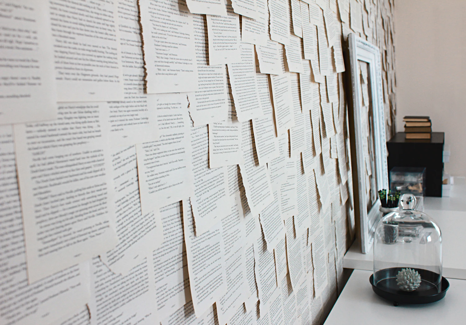 A long wall papered with hundreds of torn book pages pinned flat, a white desk running beneath it