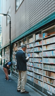A narrow alley with an outdoor bookshelf filled with books lining a brick wall. Two individuals are browsing the books; one person is intently reading a book while standing, and another is bent over examining the lower shelves. The scene conveys a quiet and contemplative atmosphere, typical of a book market or street library.