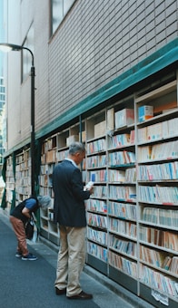 A narrow alley with an outdoor bookshelf filled with books lining a brick wall. Two individuals are browsing the books; one person is intently reading a book while standing, and another is bent over examining the lower shelves. The scene conveys a quiet and contemplative atmosphere, typical of a book market or street library.