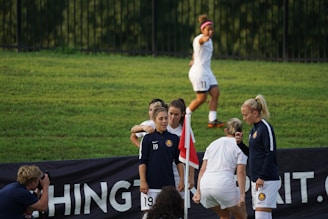 A group of female soccer players gather near a corner flag on a grassy field, with one player in a jacket and the others in team uniforms. Another player is jogging in the background, while a photographer captures the moment.