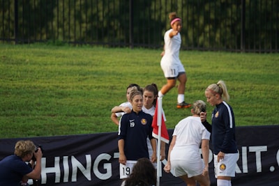 A group of female soccer players gather near a corner flag on a grassy field, with one player in a jacket and the others in team uniforms. Another player is jogging in the background, while a photographer captures the moment.