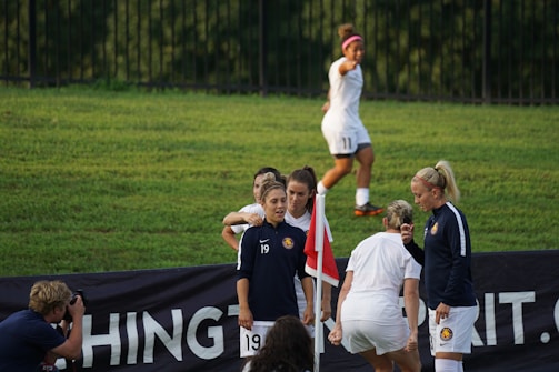 A group of female soccer players gather near a corner flag on a grassy field, with one player in a jacket and the others in team uniforms. Another player is jogging in the background, while a photographer captures the moment.
