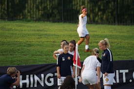 A group of female soccer players gather near a corner flag on a grassy field, with one player in a jacket and the others in team uniforms. Another player is jogging in the background, while a photographer captures the moment.