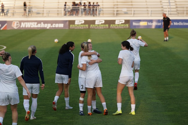 A group of women on a soccer field, some wearing white soccer jerseys and shorts. Two women in the center are embracing, while others are walking or standing nearby. A coach or official can be seen in the distance on the field. The background shows empty bleachers and advertisement banners.