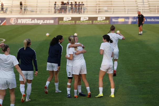 A diverse group of women celebrating together on a soccer field at sunset, wearing dark green and white jerseys.